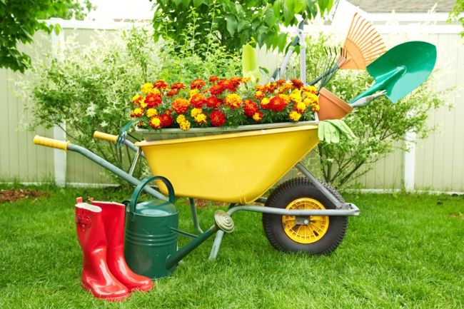 Gartenwerkzeuge, rote Gummistiefel, Gießkanne und Schubkarre mit bunten Blumen als einer abwechslungsreichen Ergänzung des ExterieursGartendeko - Ideen