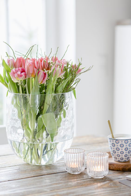 Schöne Blumenarrangements rosafarbene Tulpen im Glasgefäß auf einem Holztisch zwei Kristallgläser ein Becher