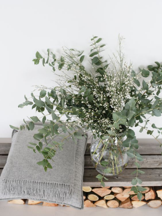 Schöne Blumenarrangements rustikales Arrangement grüne Eukalyptuszweige im Glasgefäß Brennholz daneben ein Deko Kissen